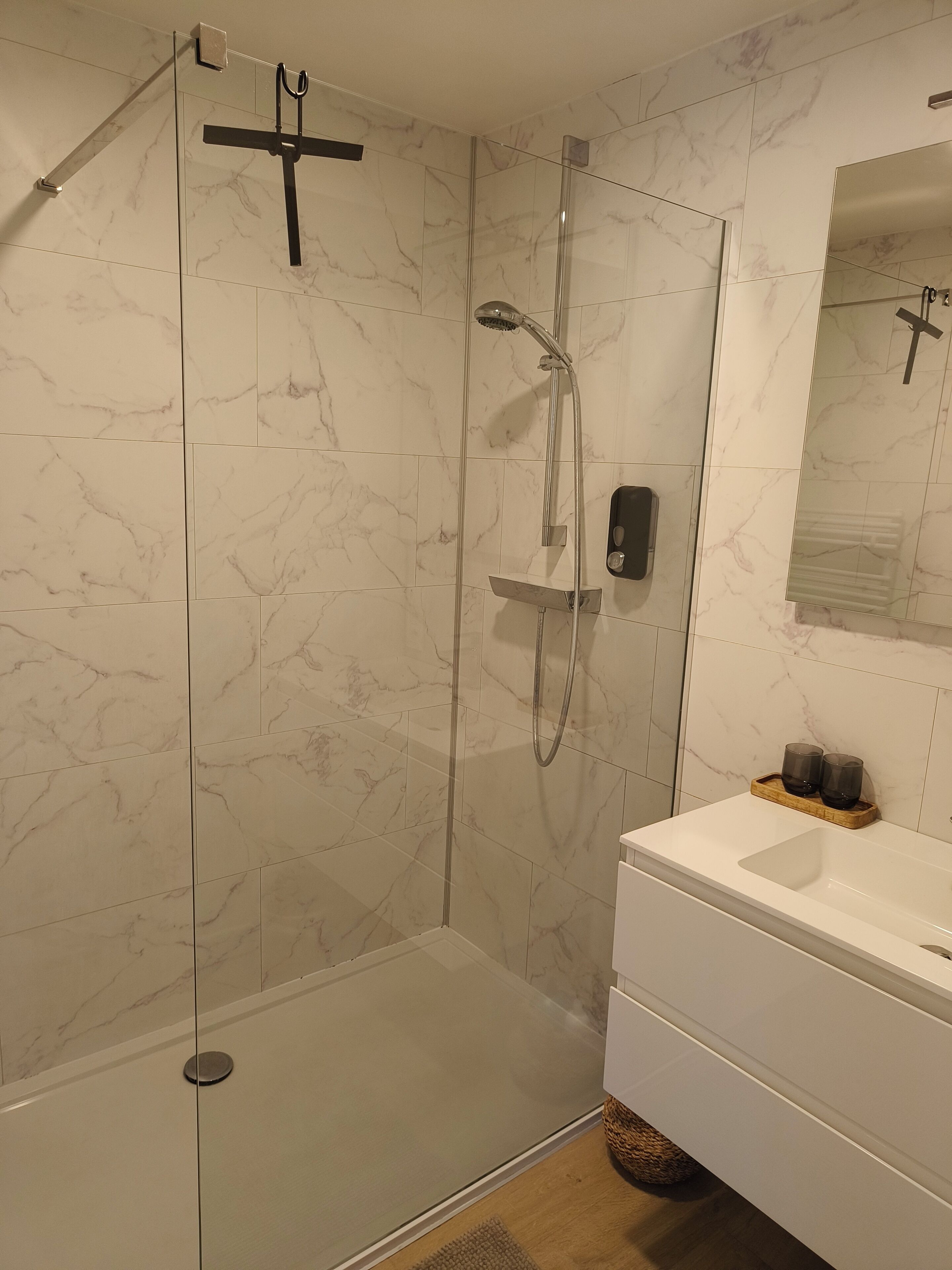 Et moderne badeværelse med en glasvæg omkring brusebadet, en hvid vask og et spejl over vasken.