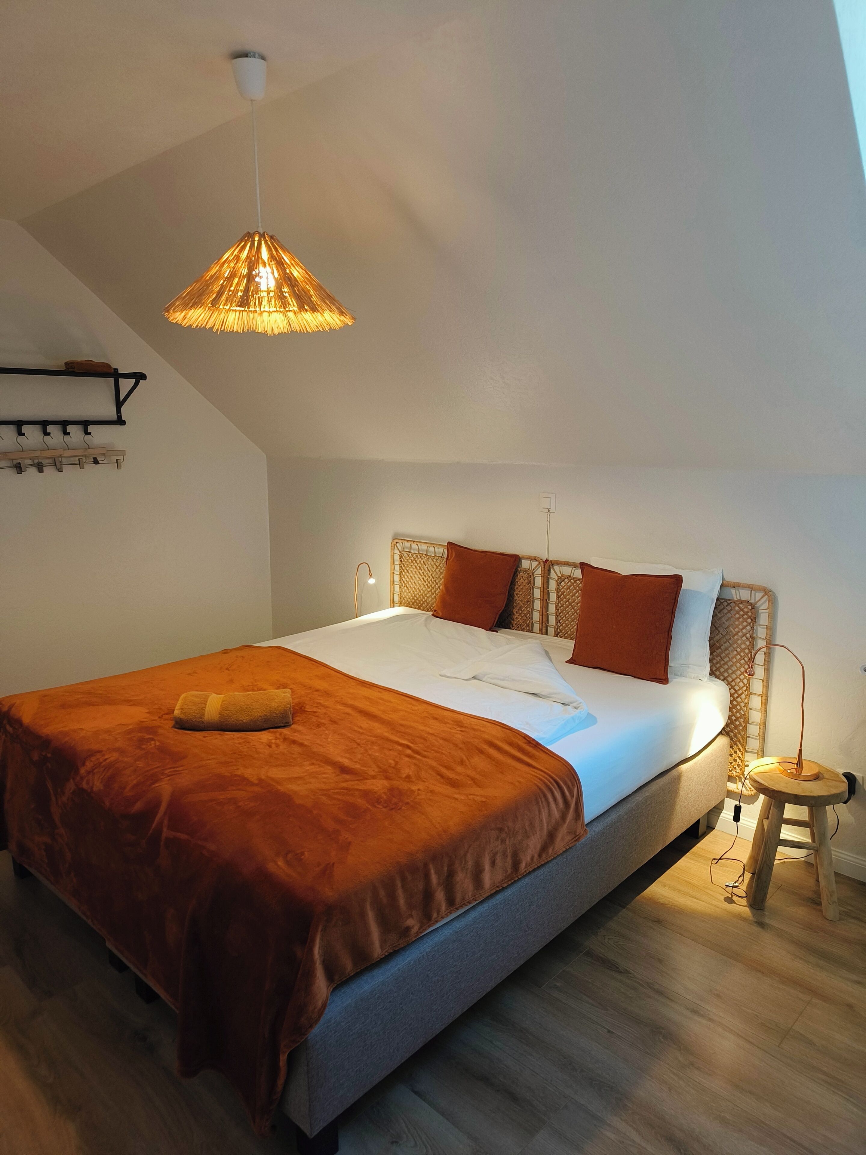 Een slaapkamer met een groot bed, een hanglamp, een houten bankje en een kapstok aan de muur.