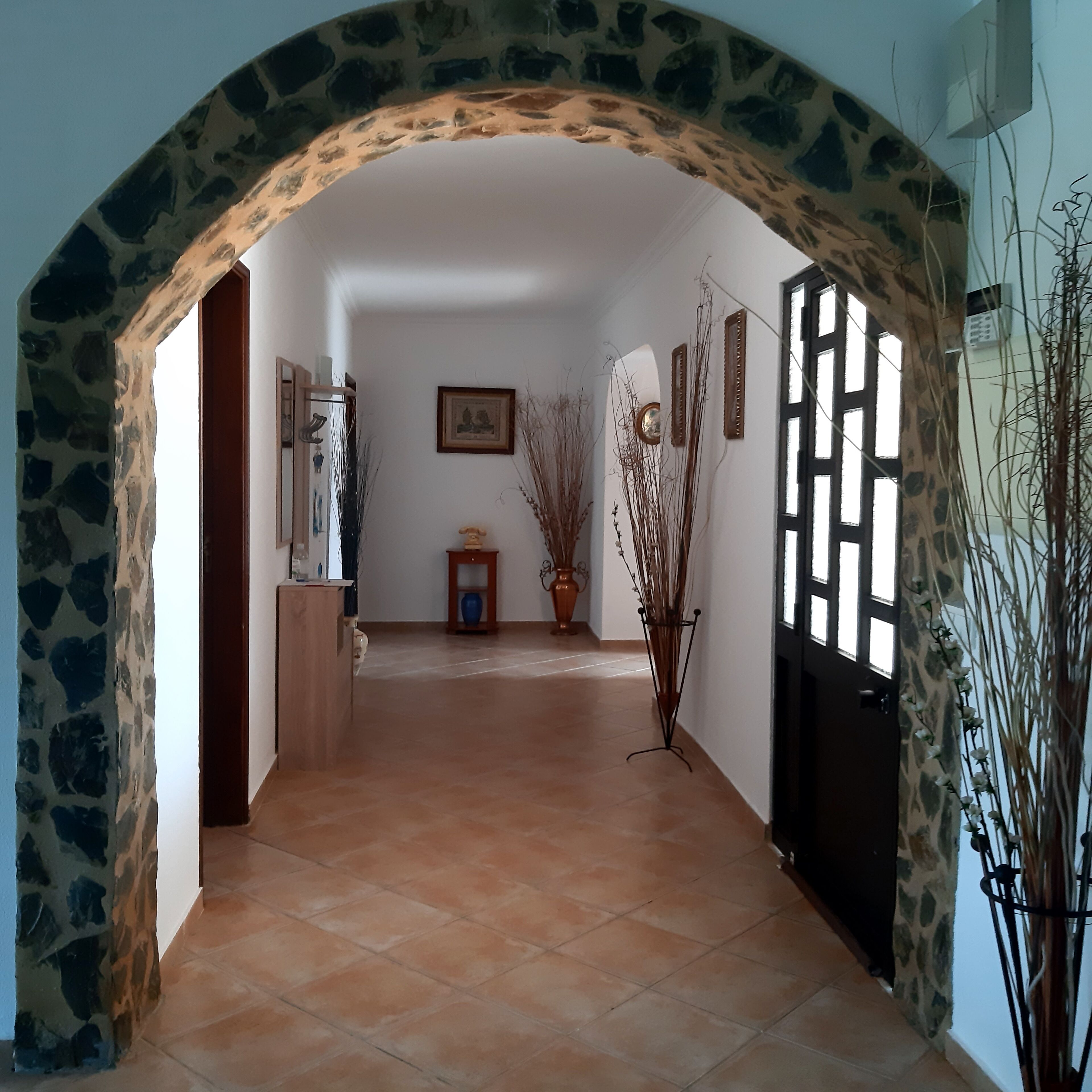 A hallway with a stone archway, a tiled floor, a wooden cabinet, a small table with a vase, and a potted plant.