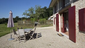 Une terrasse avec une table et des chaises, un parasol et un chemin de gravier menant à un bâtiment aux volets rouges.