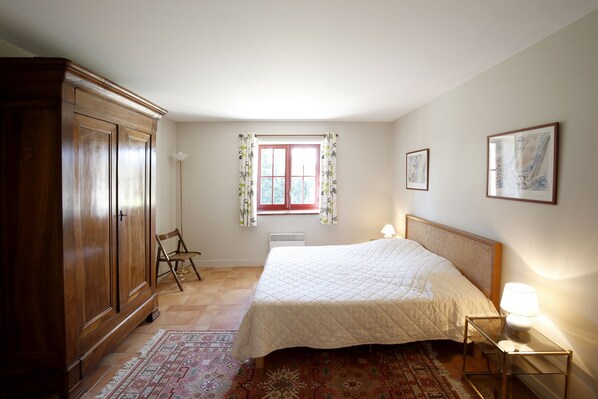 A bedroom with a bed, a nightstand, a lamp, a chair, a wardrobe, a window with curtains, and two framed pictures on the wall.