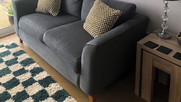 A grey sofa with two patterned cushions, a small wooden side table with a lamp, and a view of a body of water through a window with grey curtains. - Harbour-Side Apartment In Porthmadog With Stunning Views (Porthmadog)