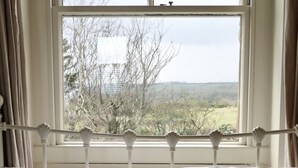 A bed with white bedding and striped pillows, a white headboard with decorative finials, and a view of trees and hills through a window with curtains. - Coastguard Cottage With Sea Views (fairlight)