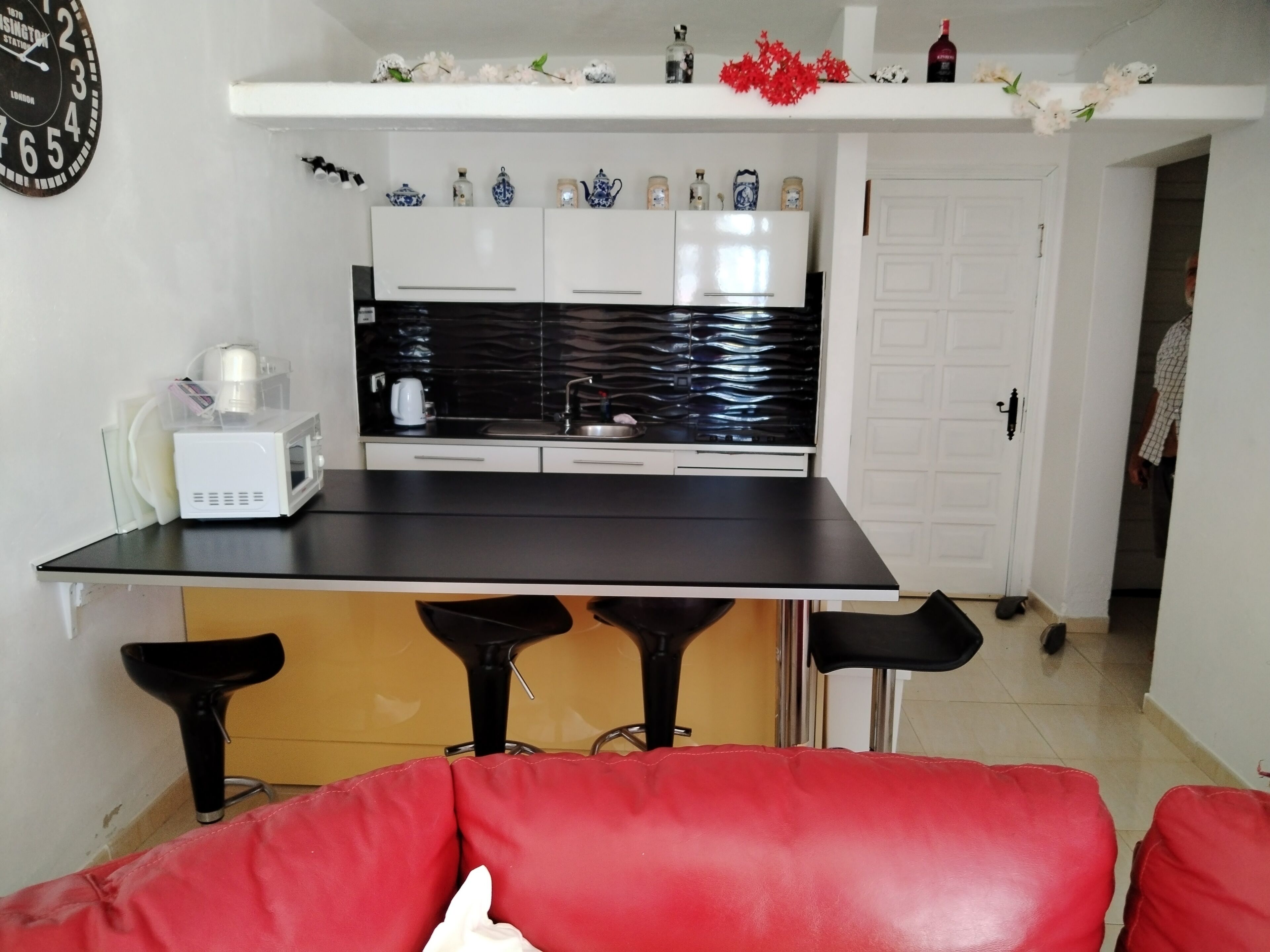 A compact kitchen with a black countertop, white cabinets, and a microwave. There are three red bar stools and a white door in the background.