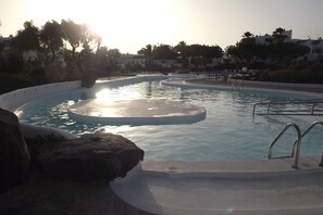 A swimming pool with a sunlit water feature, surrounded by rocks and palm trees, with buildings in the background. - Holiday apartment in a private complex with beautiful pools and gardens (Costa Teguise)