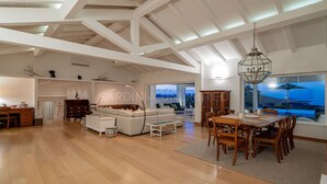 Un vaste espace de vie doté d’un haut plafond, d’un plancher en bois, d’une table à manger avec des chaises, d’un canapé et offrant une vue sur l’extérieur grâce à de grandes fenêtres.
