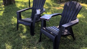 Dos sillas Adirondack con un vaso de agua y una revista sobre el césped, cerca de un árbol y una valla de madera.