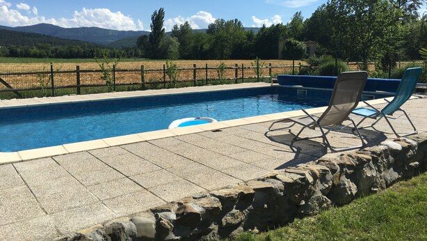 Una piscina rectangular con borde de piedra, dos sillas de descanso y vista a un campo cercado y montañas al fondo.