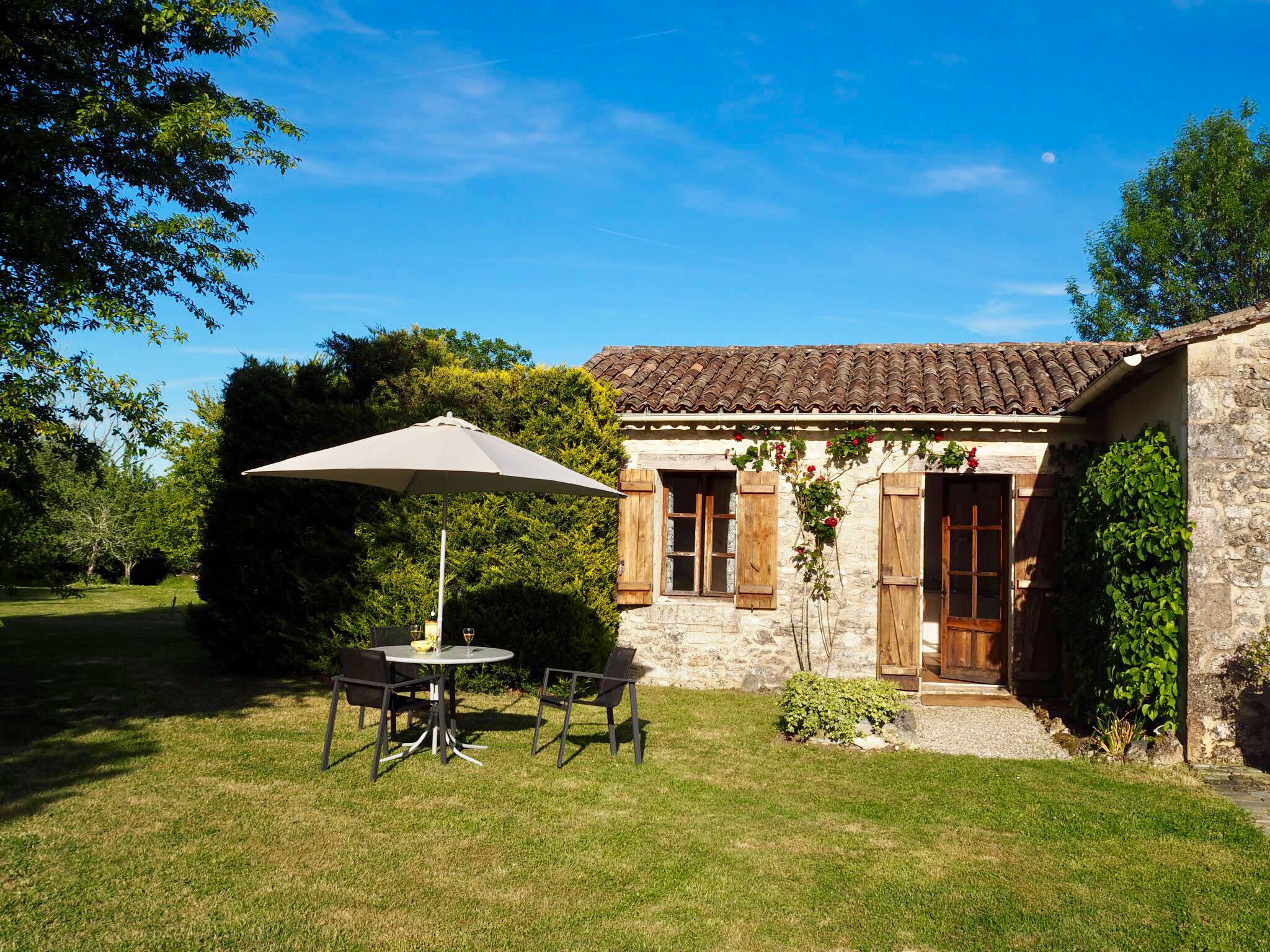 A stone building with a tiled roof, a wooden door, and a window with shutters. There is a table and chairs with an umbrella in front of the building.