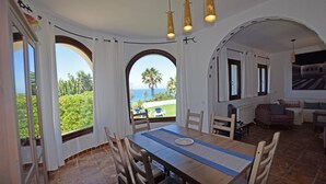 Un espace repas comprenant une table et des chaises, de grandes fenêtres avec des rideaux, et une vue sur la mer et la verdure.