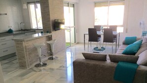 A modern living room with a sofa, a coffee table, and a TV. There is a kitchen area with a sink and a window.
