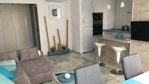 A modern living room with a sofa, a coffee table, and a dining area with chairs. There is a kitchen with a microwave, refrigerator, and a TV.