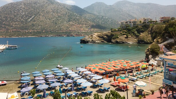 A coastal beach with rows of sun umbrellas and lounge chairs, a boat docked nearby, and a mountainous backdrop. - Sea-esta Beach House, Beachfront Residence & BBQ (Rethymno)