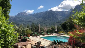 Área de piscina com espreguiçadeiras e guarda-sóis, cercada por vegetação exuberante e montanhas ao fundo.