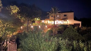 Uma casa grande de dois andares com fachada de pedra e várias janelas, rodeada por um jardim bem cuidado com diversas plantas e uma palmeira.