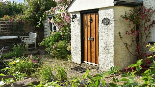 Un pequeño edificio de paredes de piedra con una puerta de madera, un banco y un jardín con diversas plantas y flores.
