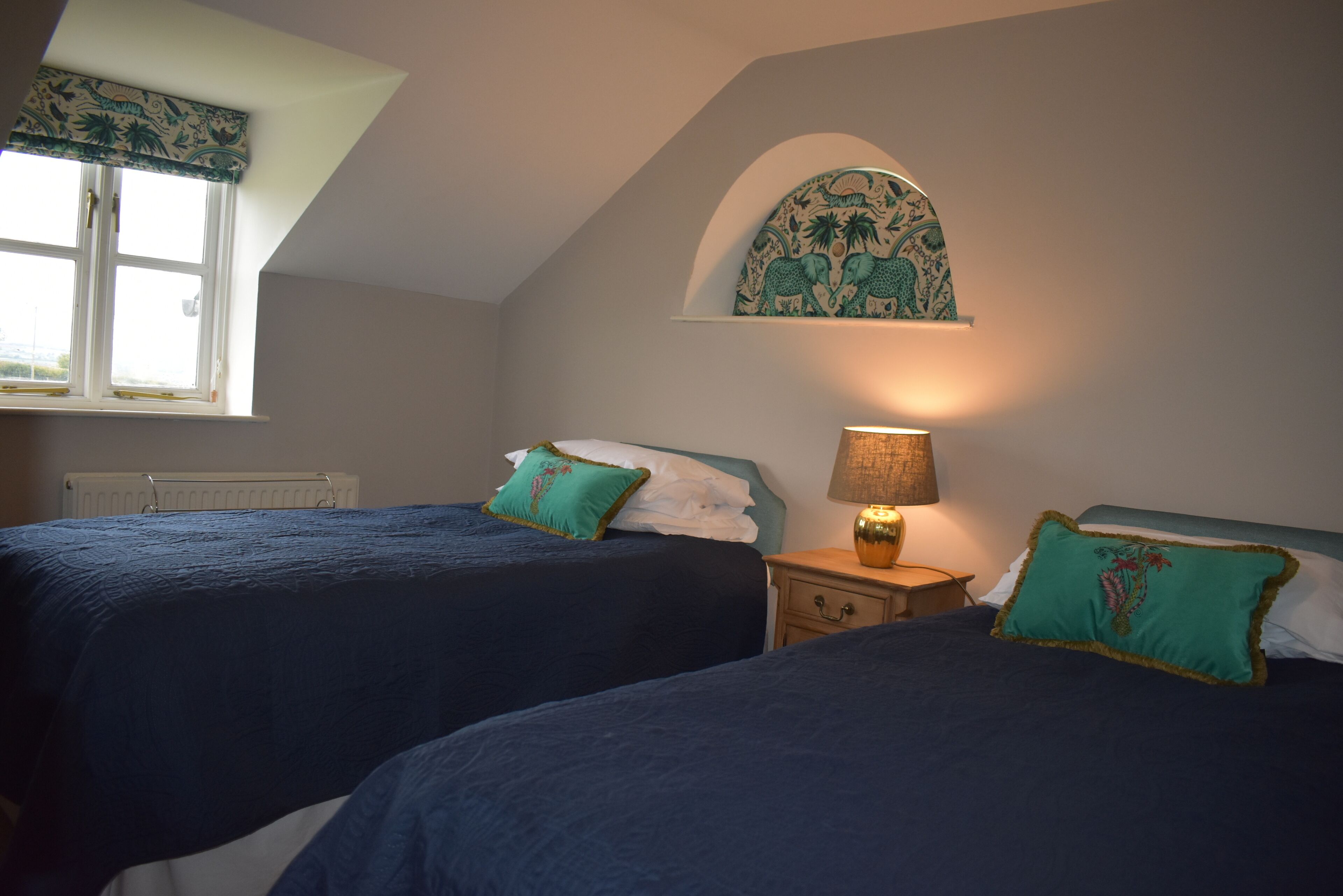 A bedroom with two beds, a nightstand with a lamp, a window with a curtain, and a decorative wall piece above the beds.