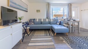A modern living room with a sectional sofa, a flat-screen TV, a dining area with a table and chairs, and a large window with curtains.