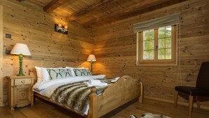 A wooden cabin bedroom with a bed, two bedside lamps, a nightstand, a chair, and a window with a view of greenery. - Chalet Jessica - La Tzoumaz, 4 Vallees (La Tzoumaz)
