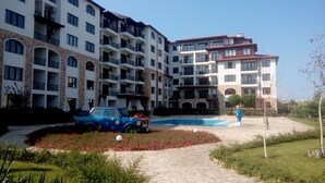 Un immeuble résidentiel à plusieurs étages, doté de balcons, d’une piscine et d’un espace jardin, avec une voiture bleue garée à proximité.