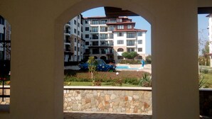 Une vue à travers une arche d’un immeuble résidentiel à plusieurs étages, avec une piscine et une voiture bleue garée devant.