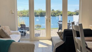 A room with a bed, a chair, a desk, and a view of a lake through large glass doors. - BOOK NOW ON LINE (Cirencester)