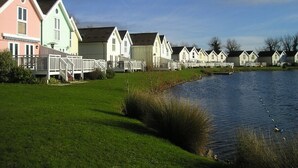 A row of colorful houses by a calm lake with a grassy foreground and a clear sky. - BOOK NOW ON LINE (Cirencester)