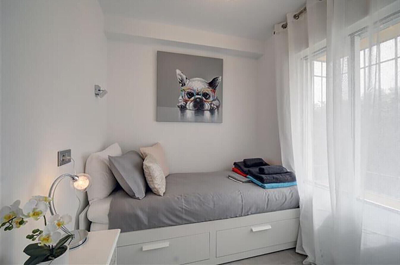A bedroom with a bed, a bedside table with a lamp, a window with curtains, and a painting on the wall.