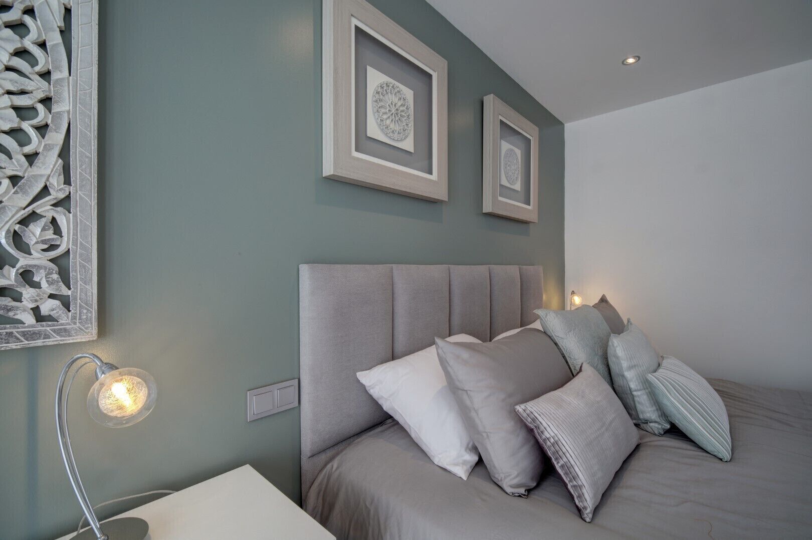 A bedroom with a bed, pillows, a nightstand, a wall-mounted light, and framed artwork.
