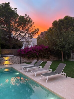 Una piscina con sillas de descanso, rodeada de vegetación y un atardecer.