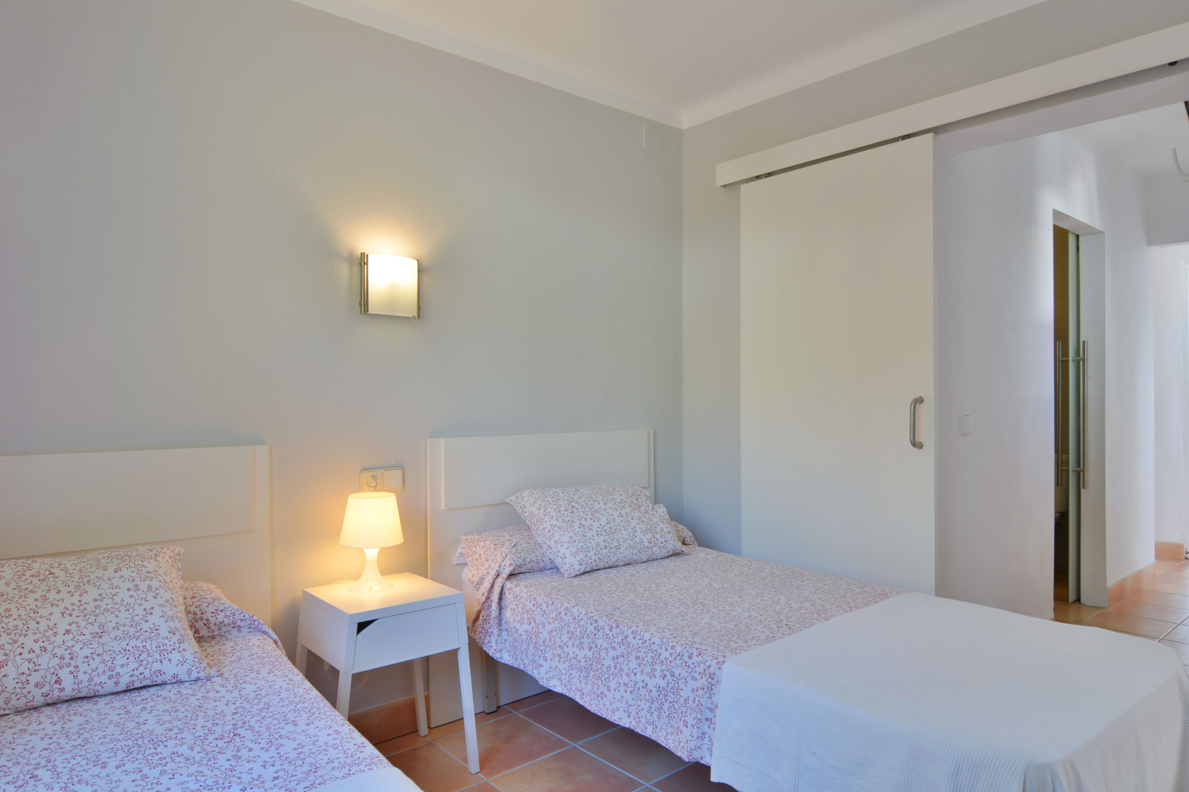A bedroom with two beds, a nightstand with a lamp, a wall-mounted light, and a sliding door closet.