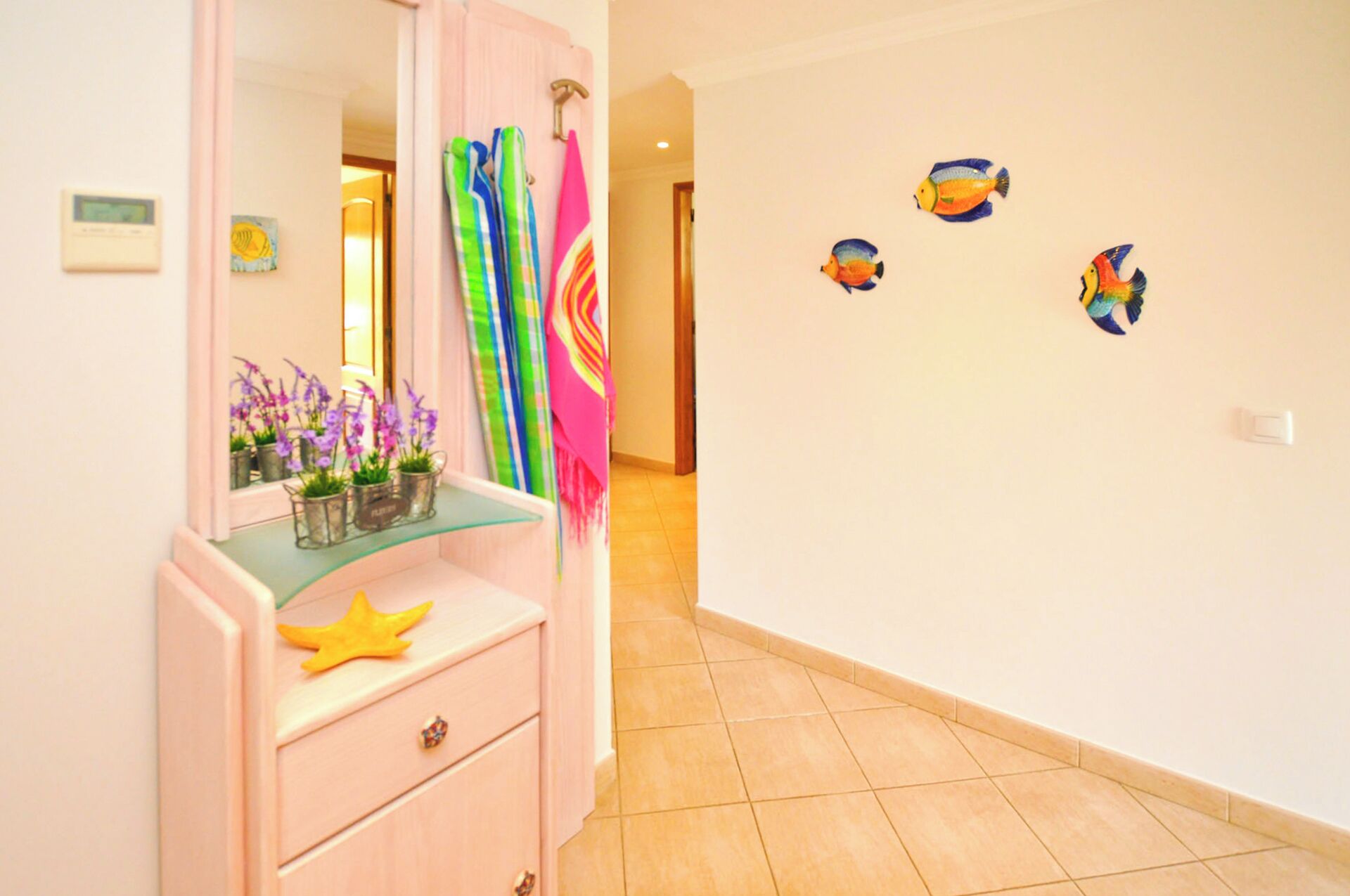 Habitación de hotel con un tocador rosa, un espejo y una estantería con flores moradas. Toallas de colores cuelgan de la pared y una puerta da a otra habitación.