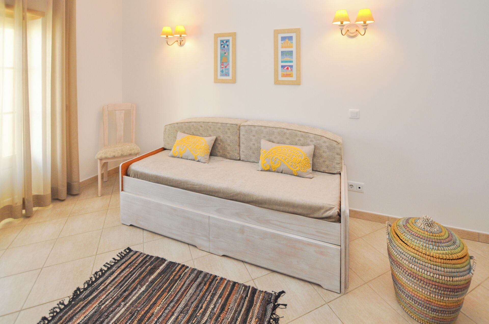 Quarto com uma cama, duas luminárias de parede, uma cadeira pequena, um banquinho tecido colorido e um tapete estampado.