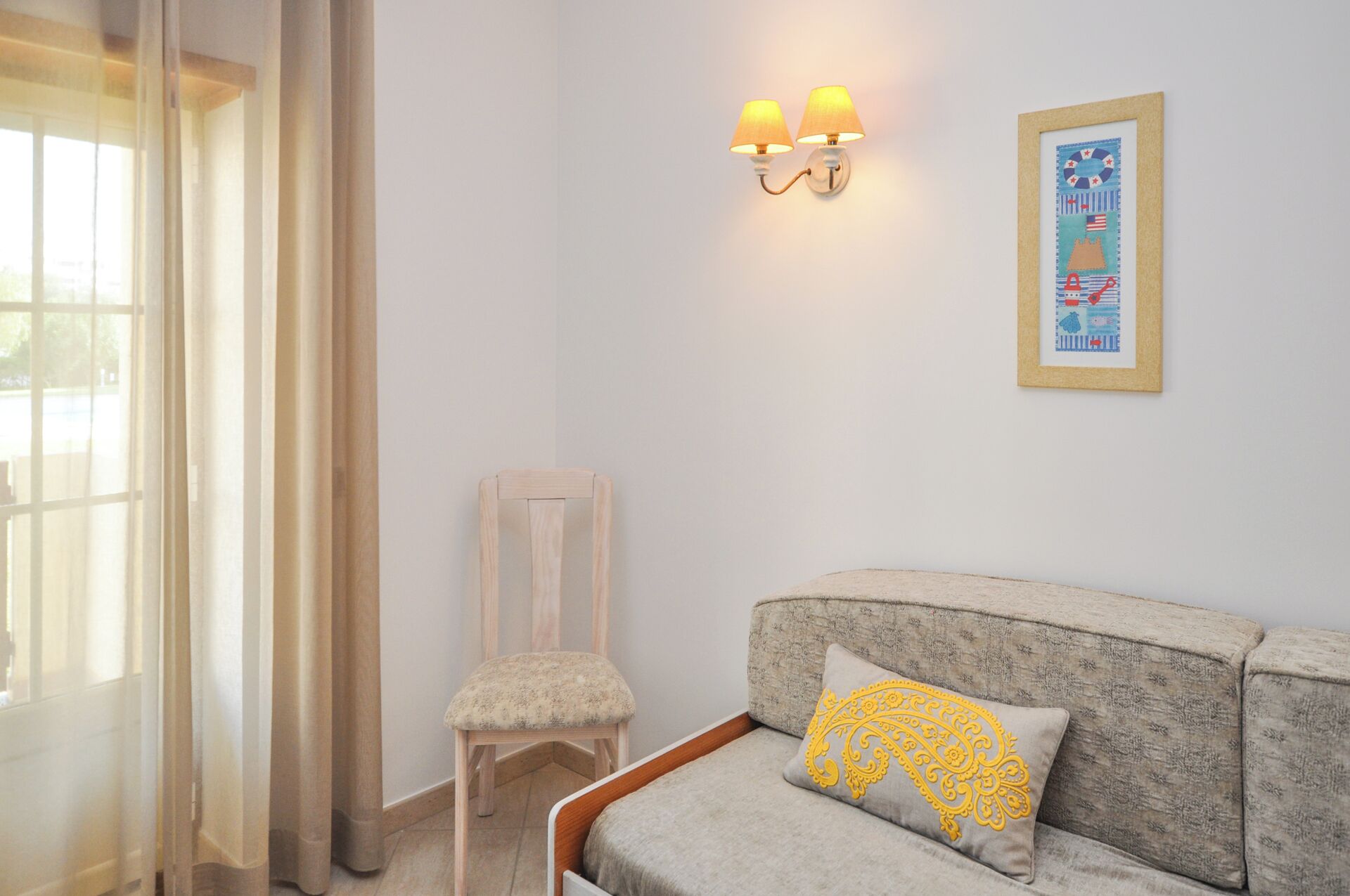 A room with a light-colored sofa, a small wooden chair, a wall-mounted lamp, and a framed picture.