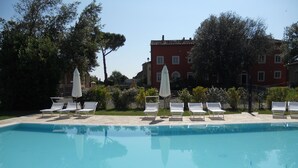 Piscina con sillas de descanso blancas y sombrillas, rodeada de árboles y edificios.