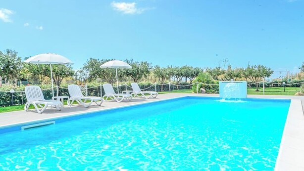 En swimmingpool med solstolar och parasoller, omgiven av grönska.