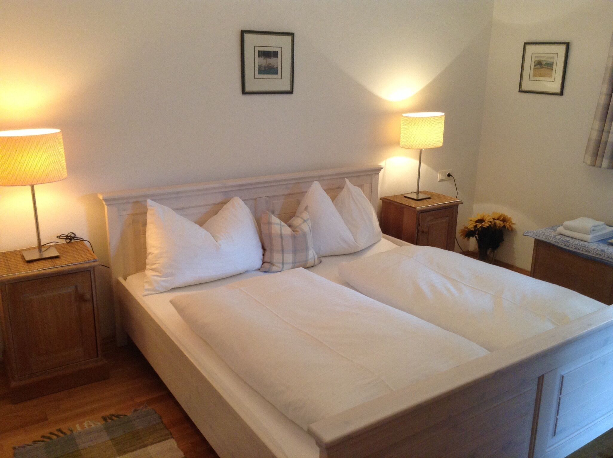 A neatly made double bed with white linens, two bedside tables with lamps, and framed pictures on the wall.