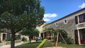 Un parque infantil con tobogán y columpios, rodeado por un edificio de piedra con contraventanas rojas y un árbol.