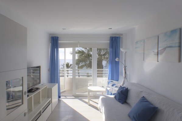 Un salón luminoso con un sofá blanco, cojines azules, un televisor sobre un mueble blanco y vistas al mar a través de amplias ventanas.