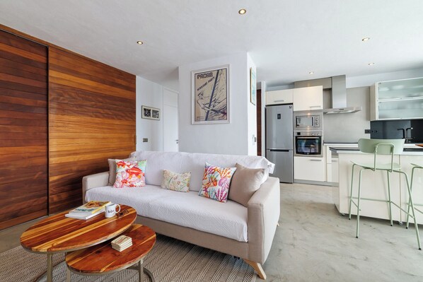 Una sala de estar moderna con sofá, una mesa de centro y una cocina con refrigerador, horno y taburetes de bar.