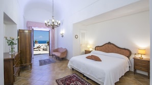 A bedroom with a large bed, bedside tables, a chandelier, a chair, and a view of the sea through an open door.