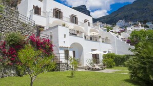 A white building with balconies and a stone wall, surrounded by greenery and mountains.