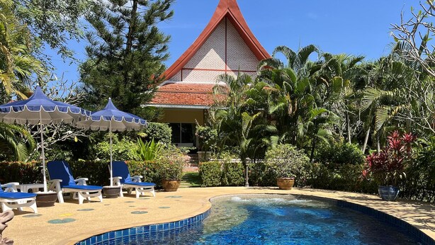 A swimming pool with blue tiles, surrounded by lounge chairs and umbrellas, with a building in the background.