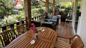 A covered outdoor seating area with wooden furniture, a table with a vase of flowers, and a view of lush greenery and a red-roofed building in the background.