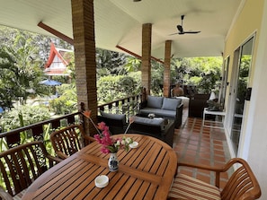 A covered outdoor seating area with wooden furniture, a table with a vase of flowers, and a view of lush greenery and a red-roofed building in the background.