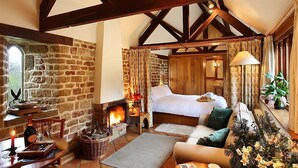 A cozy bedroom with a stone wall, wooden beams, a fireplace, a bed, and a dining area with a table set for a meal. - The cosiest and quaintest haven for couples, near Great Tew (Chipping Norton)