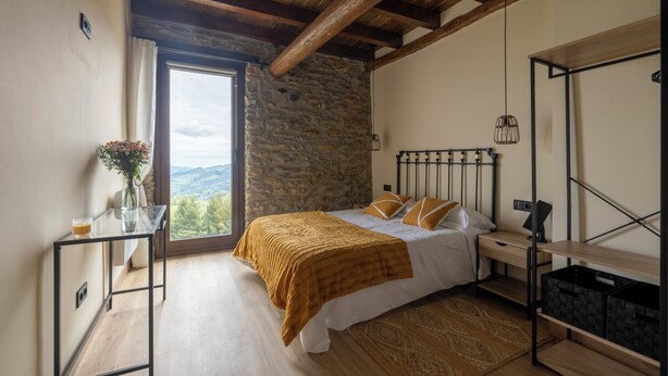 Une chambre avec un mur en pierre, des poutres en bois, un lit avec une literie blanche et des coussins de couleur moutarde, une table de chevet avec une lampe, et une vue par la fenêtre.