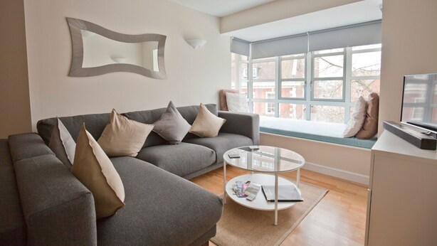 A modern living room with a grey sofa, a glass coffee table, and a large window with a view of buildings. - Toothbrush Apartments - Central Ipswich - St Peters St (Ipswich)