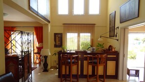 A spacious living area with a high ceiling, a dining table with chairs, a kitchen area, and large windows allowing natural light.