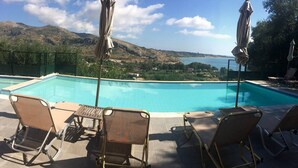 Una piscina con sillones de descanso y sombrillas, rodeada de una vista panorámica de colinas y una masa de agua.